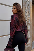 Woman wearing a patterned shirt and black pants, standing against a textured wall.