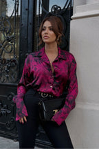 Woman wearing a vibrant pink floral blouse and black pants standing against a decorative metal gate.