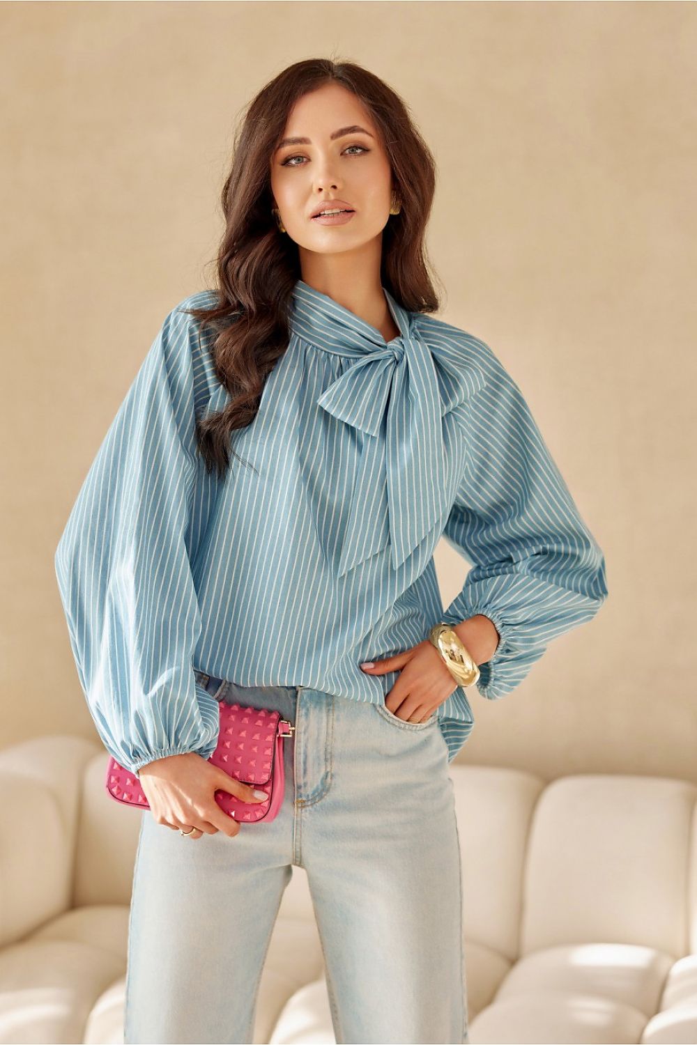 Woman wearing a blue striped blouse with a bow tie, holding a pink clutch, against a beige background.