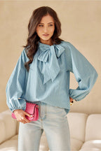 Woman wearing a light blue blouse with a bow detail, holding a pink handbag against a beige background.