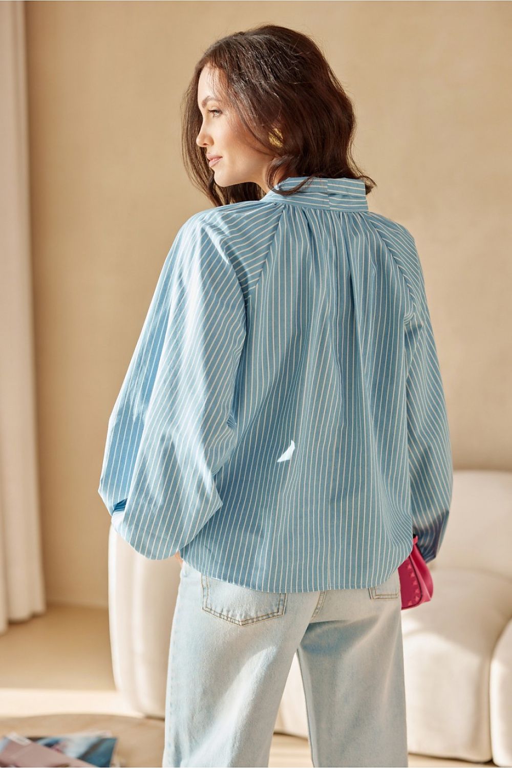 Woman wearing a light blue pleated blouse in a room with beige walls and a white couch.