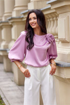 Woman wearing a pink blouse with a floral detail and white pants standing against a beige building.