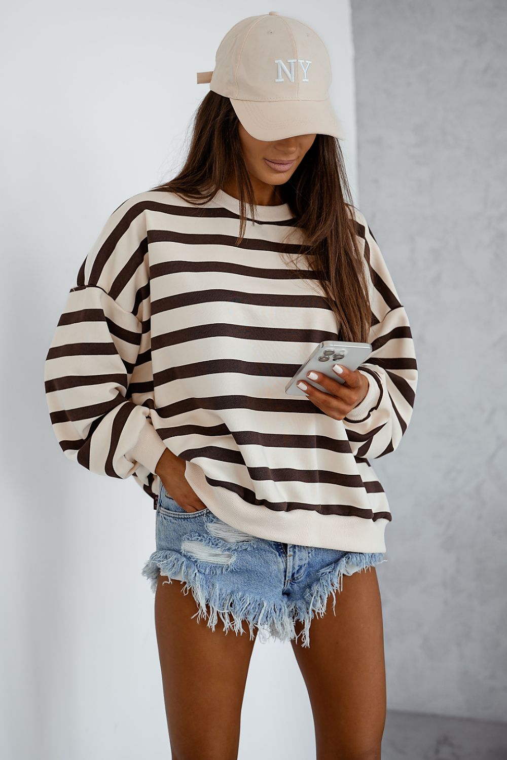 Person wearing a striped sweater, denim shorts, and a beige cap with 'NY' on a plain background