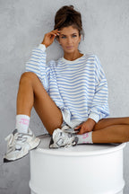 Woman wearing a blue and white striped sweater sitting on a white stool against a gray wall.
