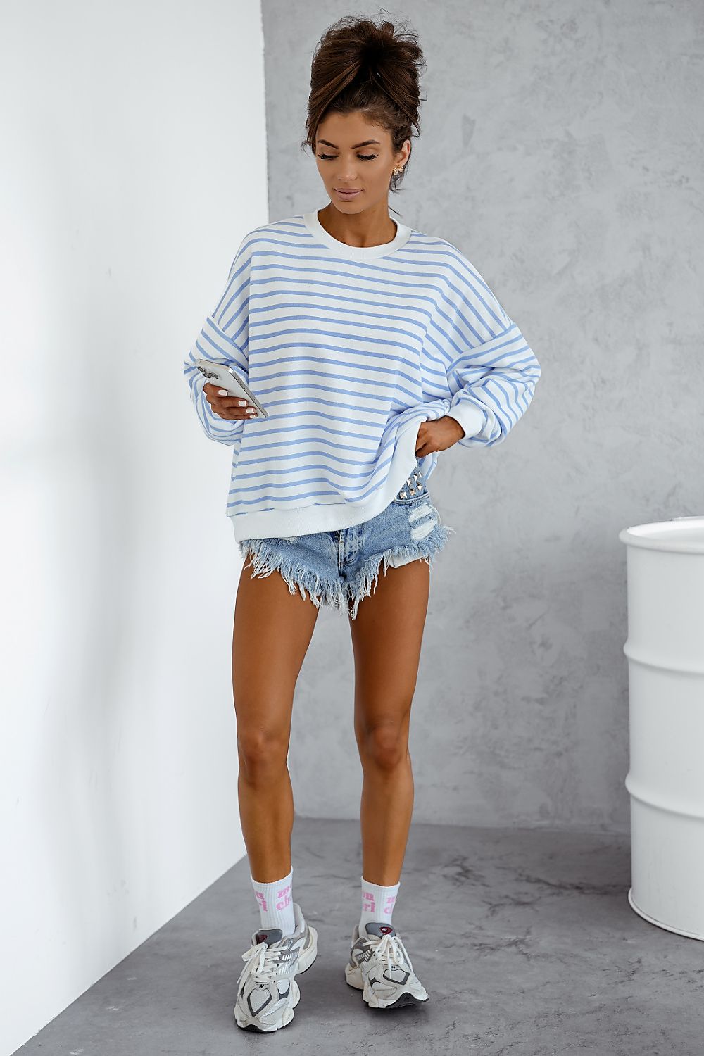 Woman wearing a blue and white striped sweater with denim shorts against a gray wall.