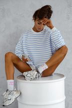Woman sitting on a white stool wearing a striped sweater and sneakers, holding a phone.