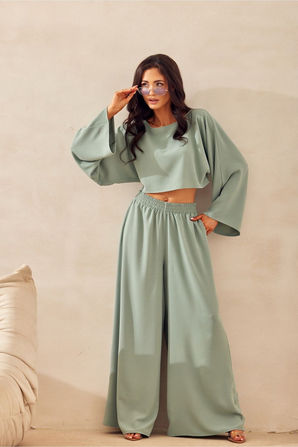 Woman wearing a light green outfit with wide sleeves and high-waisted pants against a beige background.