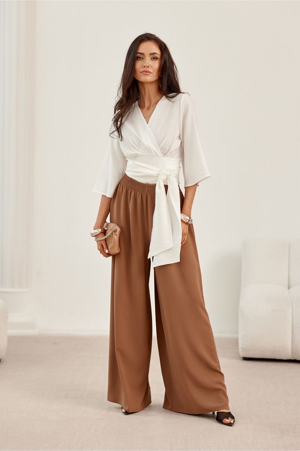 Woman wearing a white blouse and brown wide-leg pants in a minimalistic room.