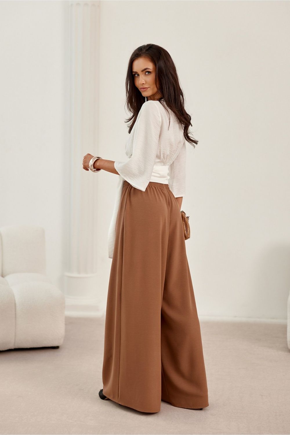 Woman wearing a white blouse and brown wide-leg pants in a minimalistic room.