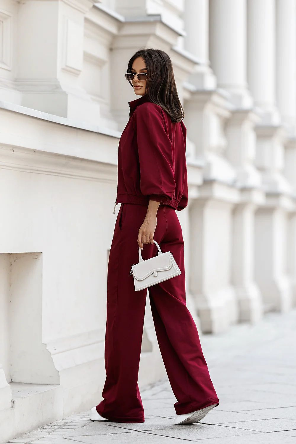 Woman in burgundy outfit holding a white handbag against a white architectural background
