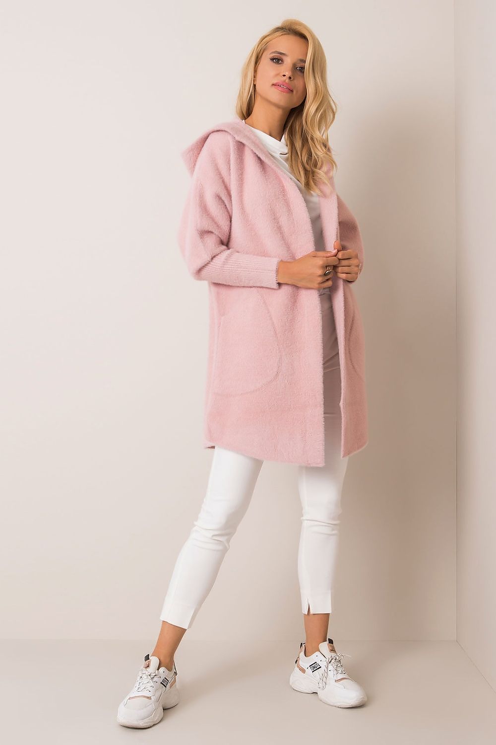 Woman wearing a pink hooded coat with white pants and sneakers on a plain background