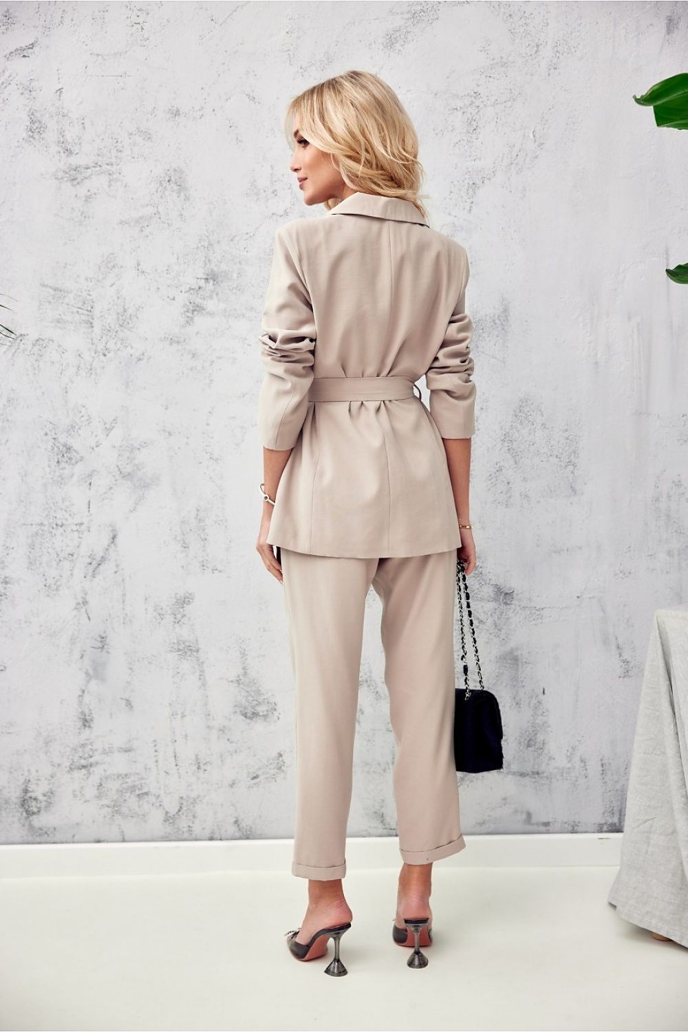 Woman wearing a beige trench coat and pants against a light gray wall.