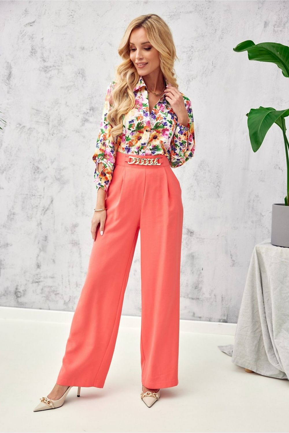 Woman wearing a colorful floral blouse and coral pants in a minimalistic room.