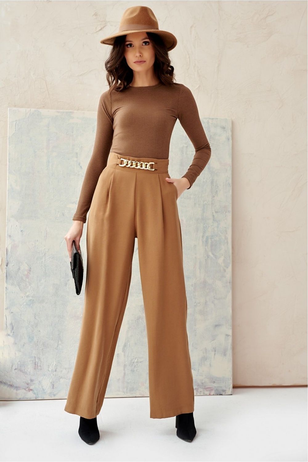 Woman wearing a brown outfit with a hat and belt, standing against a neutral background.