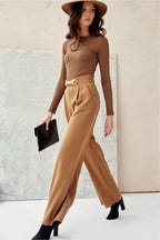 Woman in a brown outfit with wide-leg pants and a hat, holding a clutch against a neutral background.