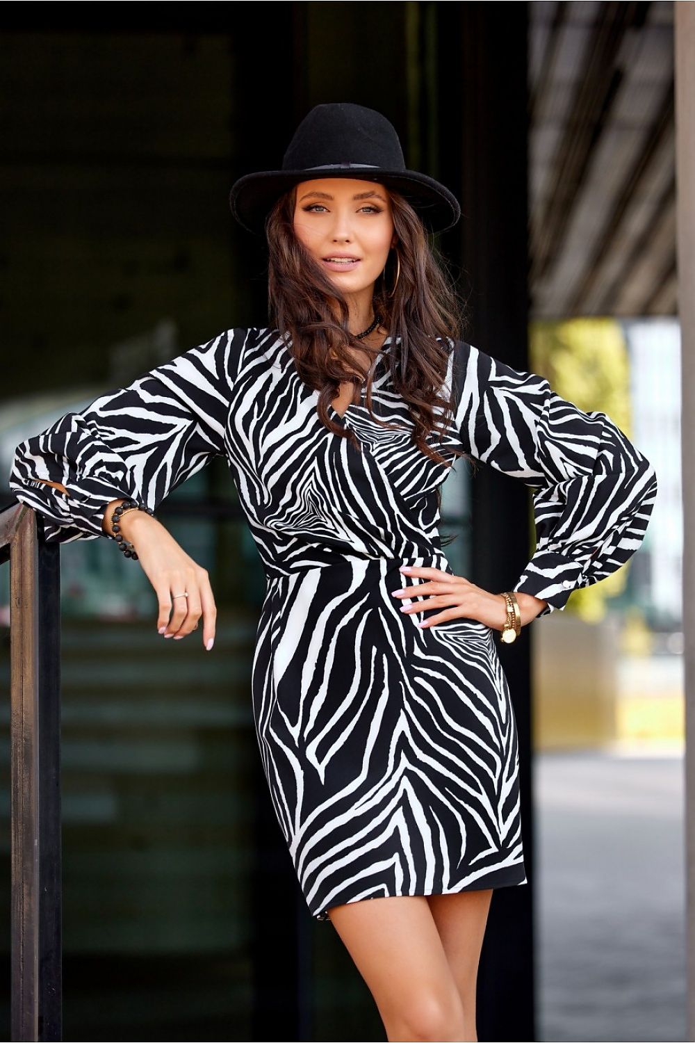 Woman wearing a zebra print dress and black hat leaning against a railing.