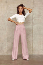 Woman wearing a white top and pink pants against a neutral wall.