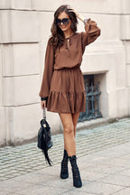 Woman wearing a brown dress with black boots and sunglasses, standing against a stone wall.