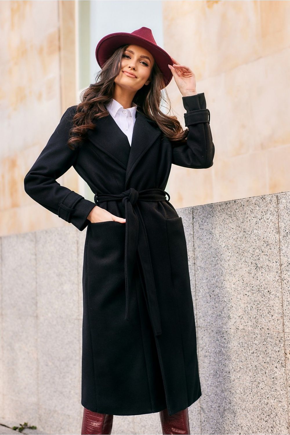 Woman wearing a black coat and red hat standing against a stone wall.