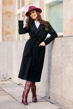 Woman wearing a black coat, white shirt, red hat, and red boots standing against a building.