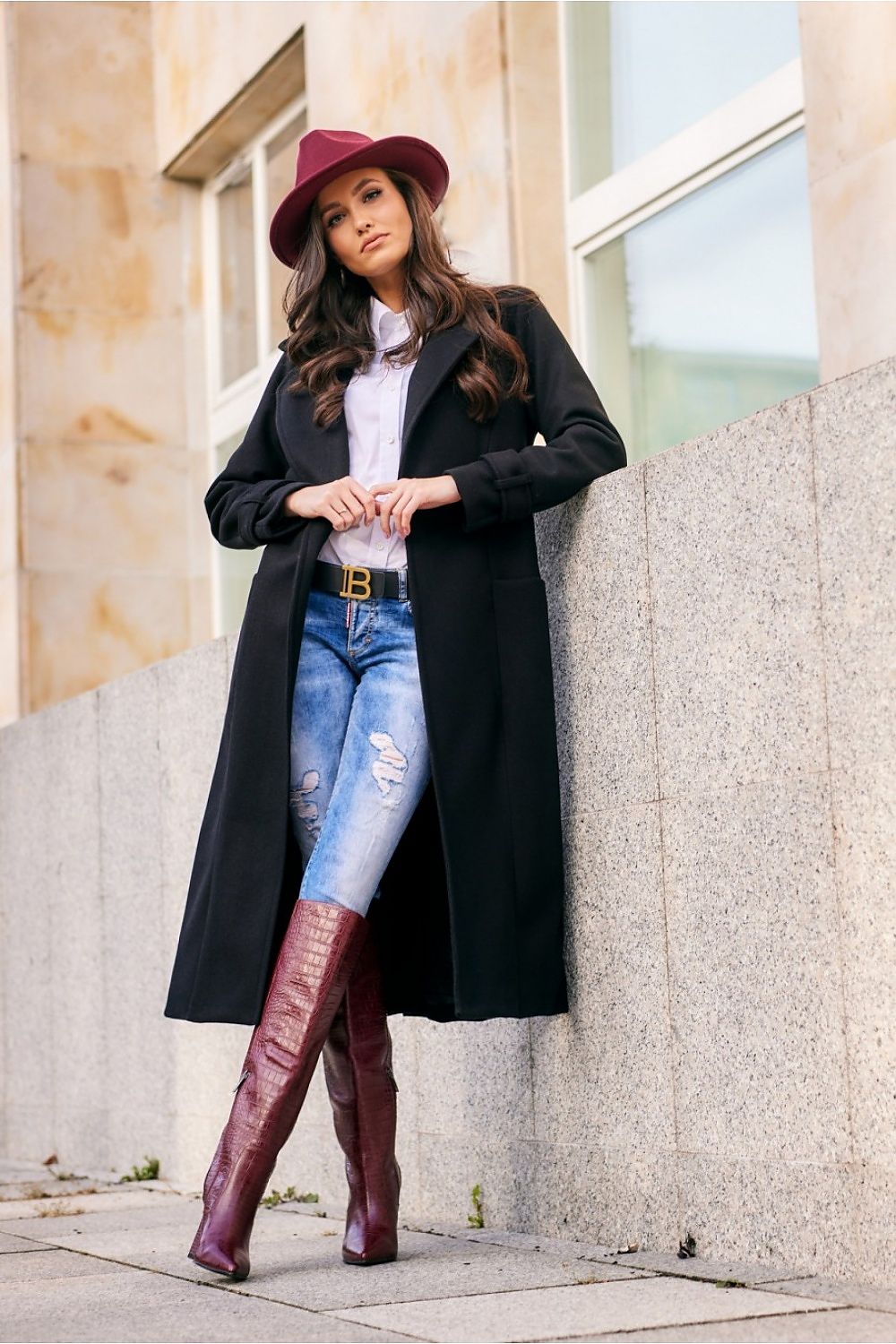 Woman wearing a black coat, white shirt, blue jeans, and maroon boots against a building.