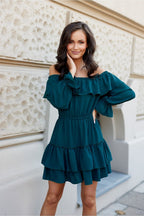 Woman wearing a teal off-shoulder dress standing against a light-colored wall.
