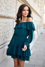 Woman wearing a teal off-shoulder dress standing against a light-colored wall.