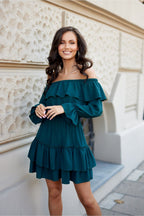 Woman wearing a teal off-shoulder dress standing against a light-colored wall.