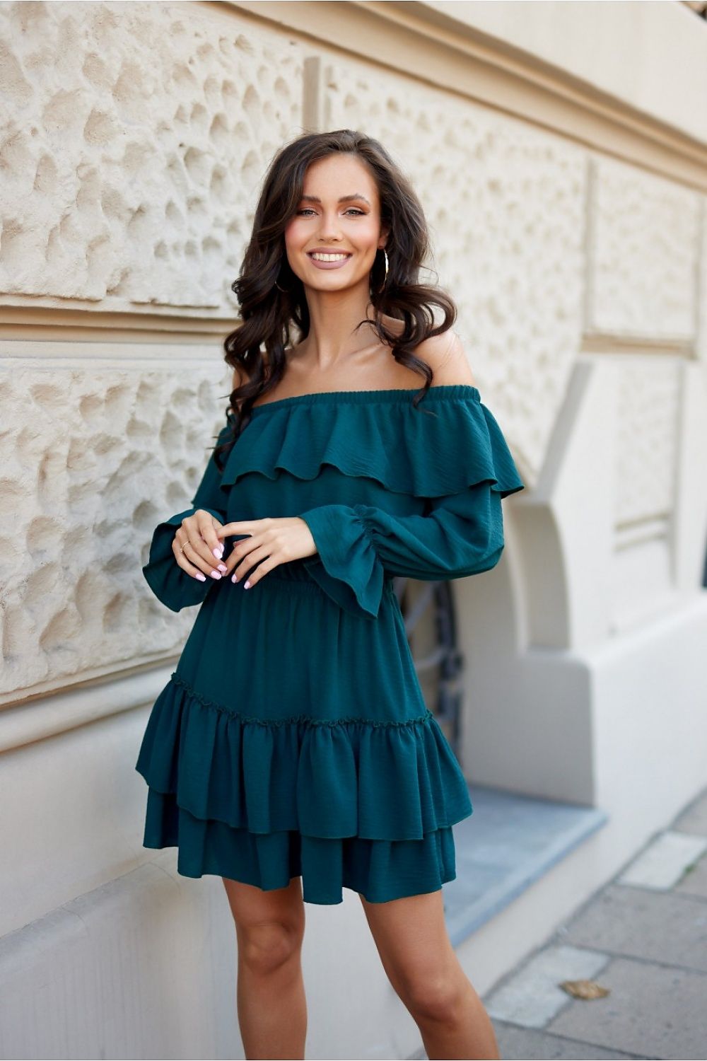 Woman wearing a teal off-shoulder dress standing against a light-colored wall.