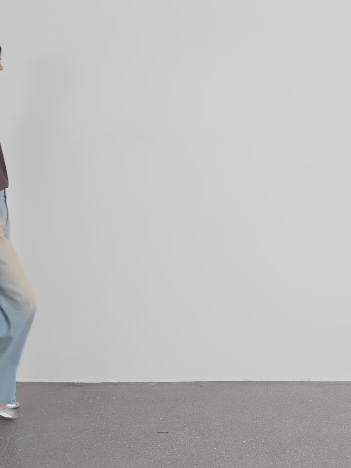 Person wearing a brown top and light blue pants walking on a gray floor with a white wall background